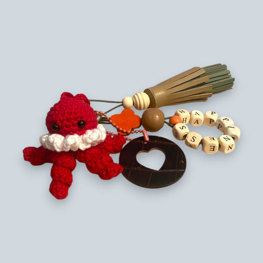 Bijou de sac en cuir avec quatre breloques fixées à un mousqueton doré ovale : un pompon beige et vert tendre avec une perle en bois, une breloque en perles de bois formant le mot « HAPPINESS », une breloque ronde en cuir marron ajourée d’un cœur, et une breloque en crochet faite à la main de couleur rouge.