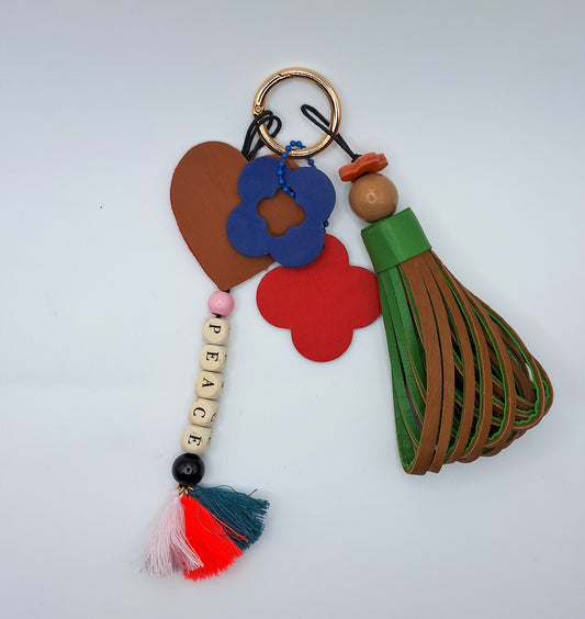 Bijou de sac en cuir et perles de bois multicolores avec anneau mousqueton doré, composé de quatre breloques : un pompon bicolore vert et marron avec une perle ronde en bois beige et un petit trèfle en cuir orange, une breloque en cuir vert et bleu en forme de trèfle évidé au centre, une breloque en cuir rose et rouge en forme de trèfle marquée M, et une breloque en perles de bois formant le mot « PEACE » avec trois pompons en fils de coton et un cœur en cuir rouge et marron au-dessus.