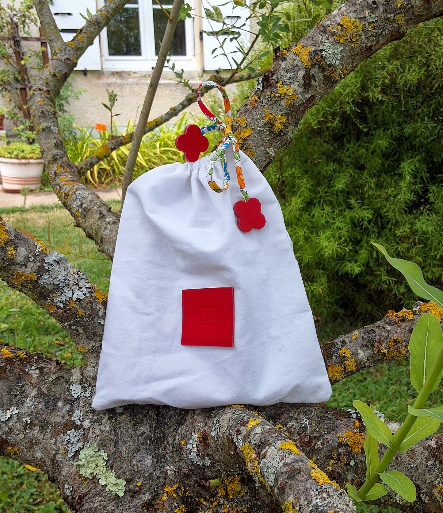 Pochon de rangement blanc avec au centre une pièce de cuir carré rouge embossé avec le M de la marque et des cordons liberty rouge disposé sur un arbre dans un jardin.