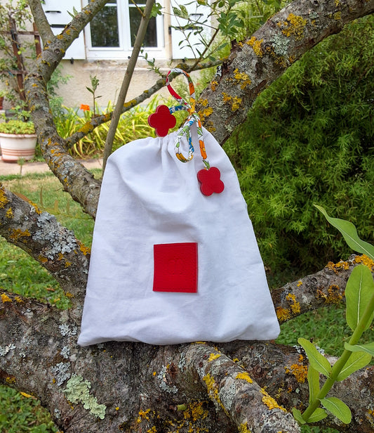 Pochon de rangement blanc avec au centre une pièce de cuir carré rouge embossé avec le M de la marque et des cordons liberty rouge disposé sur un arbre dans un jardin.