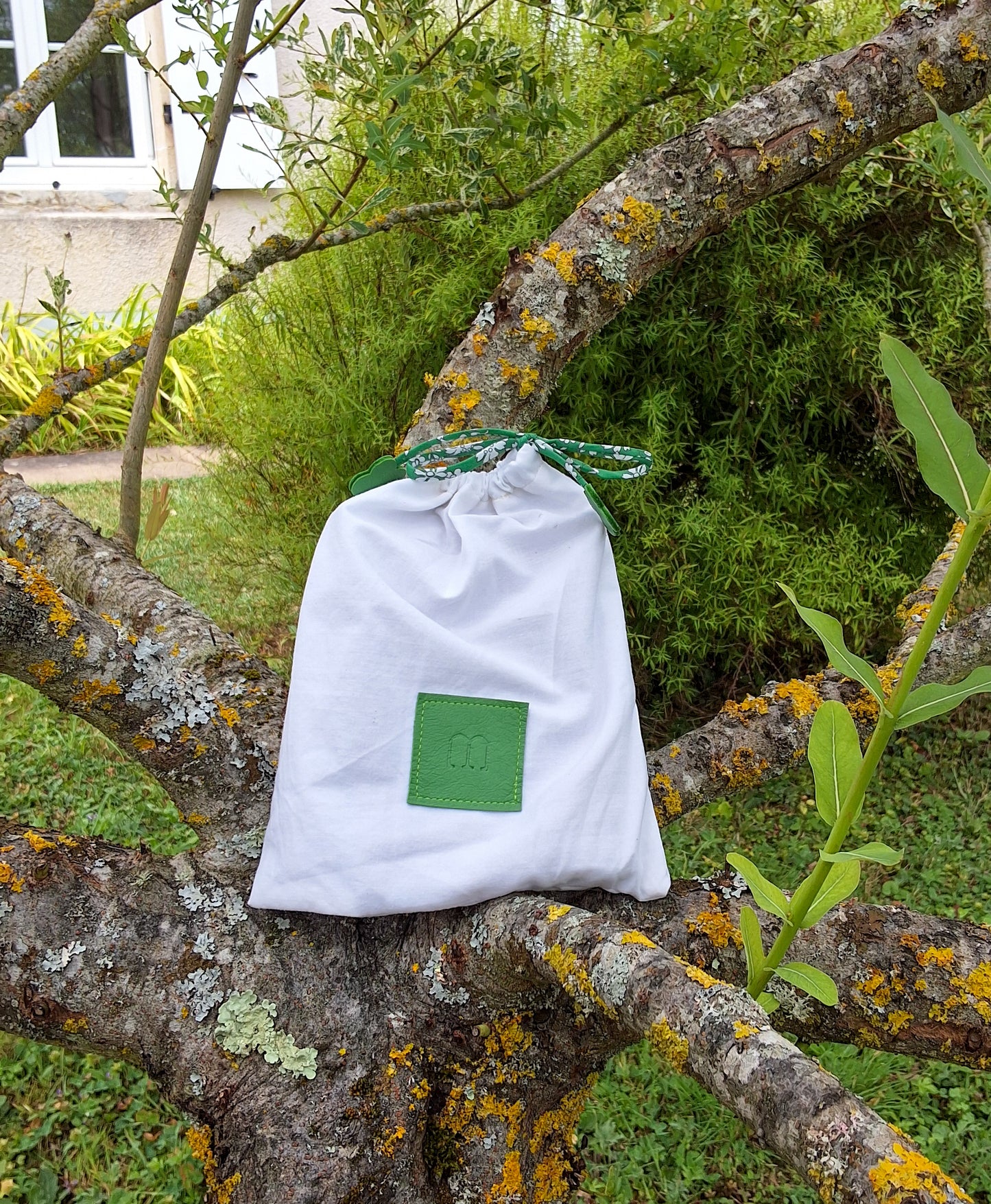 Pochon de rangement blanc avec au centre une pièce de cuir carré vert embossé avec le M de la marque et des cordons liberty vert disposé sur un arbre dans un jardin.