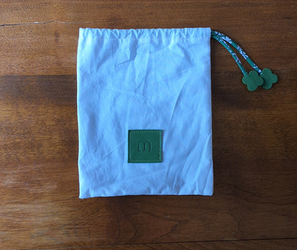 Pochon de rangement blanc avec en bas au centre une pièce de cuir carré vert embossé avec le M de la marque et des cordons liberty vert disposé à plat sur une table en bois.