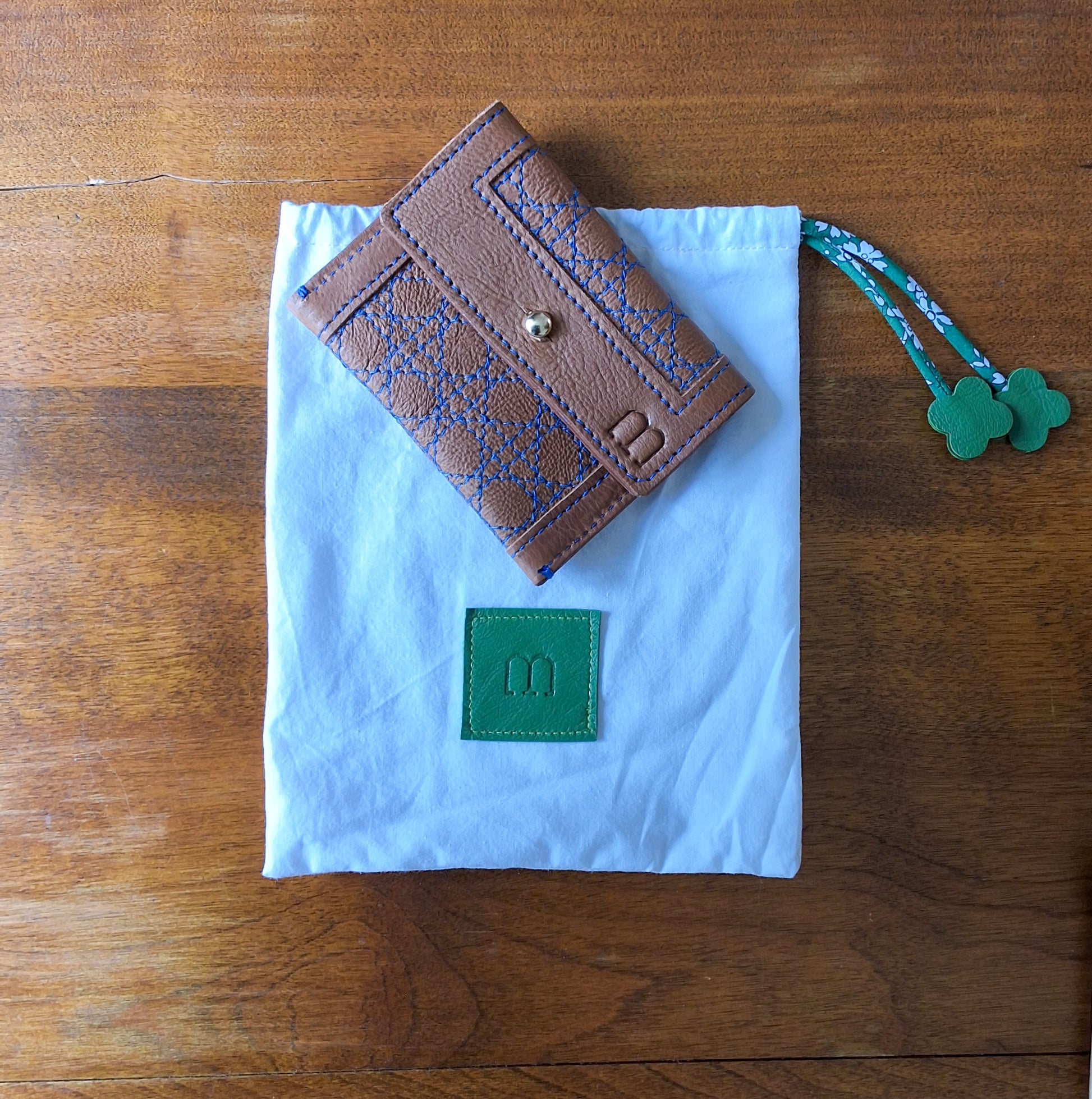 Porte cartes camel surpiqûres bleu et pochon de rangement blanc avec en bas au centre une pièce de cuir carré verte embossée avec le M de la marque et des cordons liberty vert disposés à plat sur une table en bois.