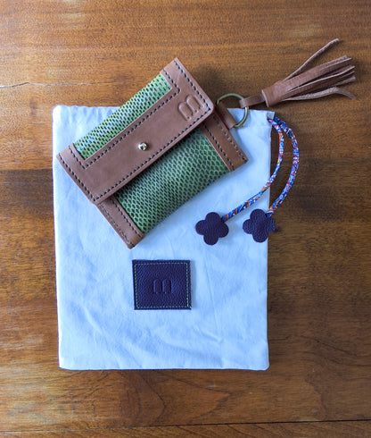 Porte cartes camel et serpent d'eau vert et pochon de rangement blanc avec en bas au centre une pièce de cuir carré violette embossée avec le M de la marque et des cordons liberty vert disposés à plat sur une table en bois.