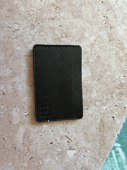 le porte carte en cuir noir saffiano est présenté à plat de face sur la pierre au bord d'une piscine. 