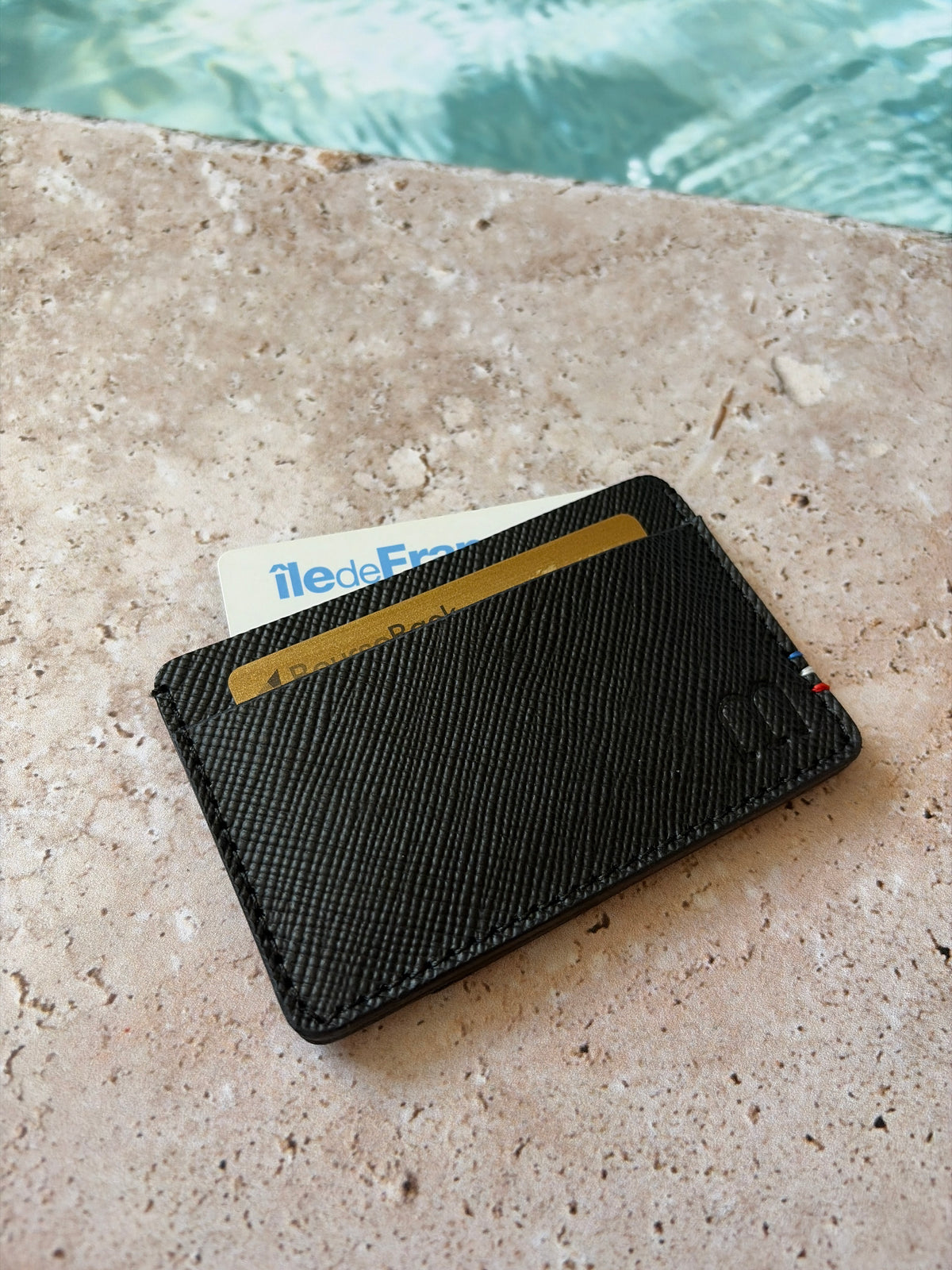 le porte carte en cuir noir saffiano est présenté les poches garnies de cartes de face sur de la pierre en bord de piscine. 