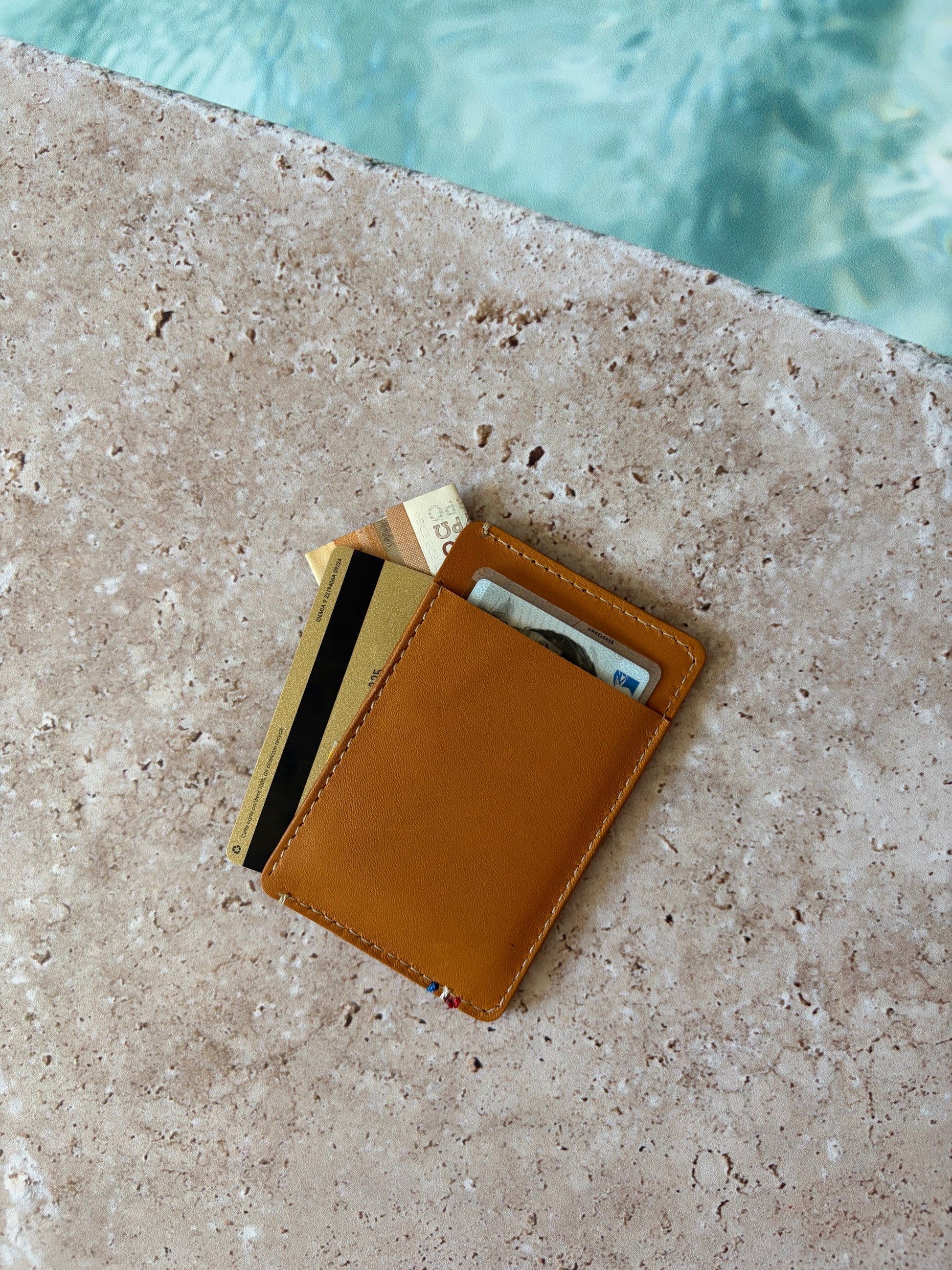 le porte carte est vu de dos posé sur de la pierre au bord d'une piscine. es poches du porte carte sont garnies. 