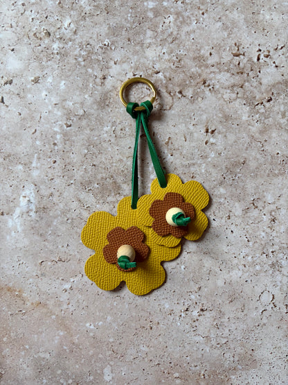 le porte clefs en cuir en forme de fleures jaune et marron est présenté à plat sur un fon den travertin. 