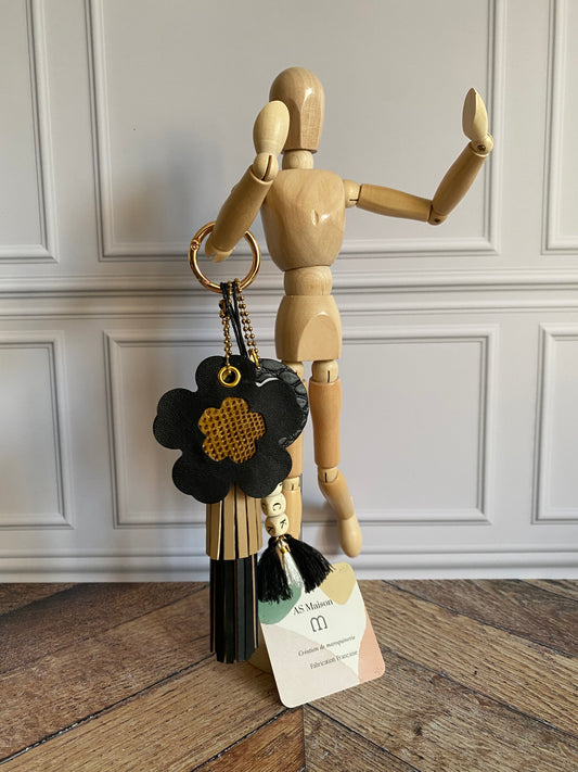 Bijou de sac avec quatre breloques en cuir fixées à un mousqueton doré : un pompon noir et beige avec deux perles en bois, une breloque ronde en cuir noir effet crocodile avec un trèfle, une breloque en forme de fleur avec un cœur en peau de serpent et un M au dos, et une breloque en perles de bois formant le mot « ROCK ».