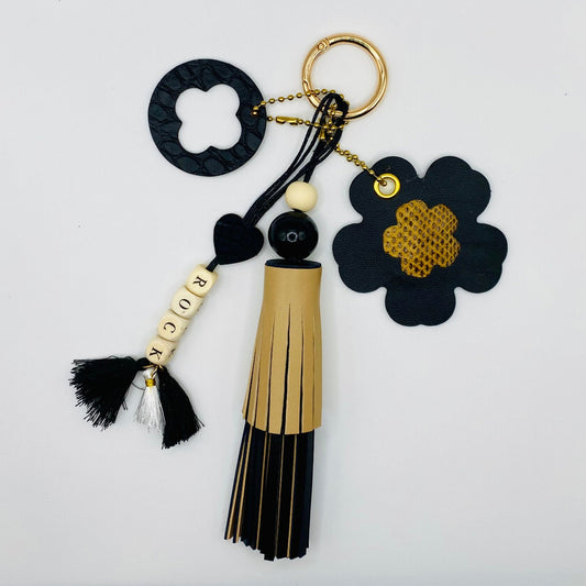 Bijou de sac avec quatre breloques en cuir fixées à un mousqueton doré : un pompon noir et beige avec deux perles en bois, une breloque ronde en cuir noir effet crocodile avec un trèfle, une breloque en forme de fleur avec un cœur en peau de serpent et un M au dos, et une breloque en perles de bois formant le mot « ROCK ».