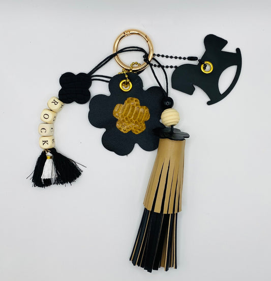 Bijou de sac avec quatre breloques en cuir fixées à un mousqueton doré : un pompon noir et beige avec deux perles en bois, une breloque en cuir en forme de cheval à bascule, une breloque en forme de fleur avec un cœur en peau de serpent et un M au dos, et une breloque en perles de bois formant le mot « ROCK ».