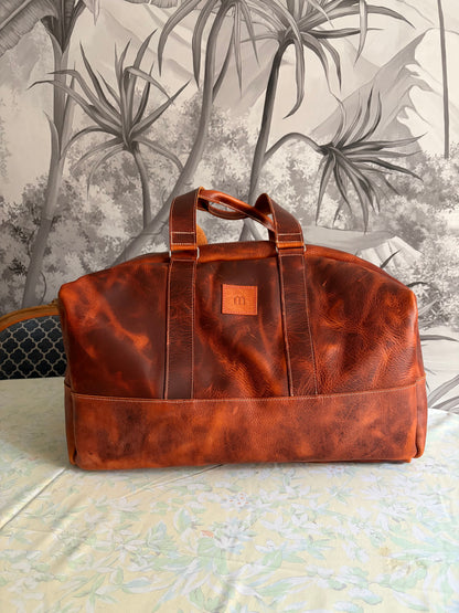 photo du sac de voyage Marius réalisé en cuir wax tannage végétal posé sur une table en bois avec en arrière plan un papier peint panoramique gris 