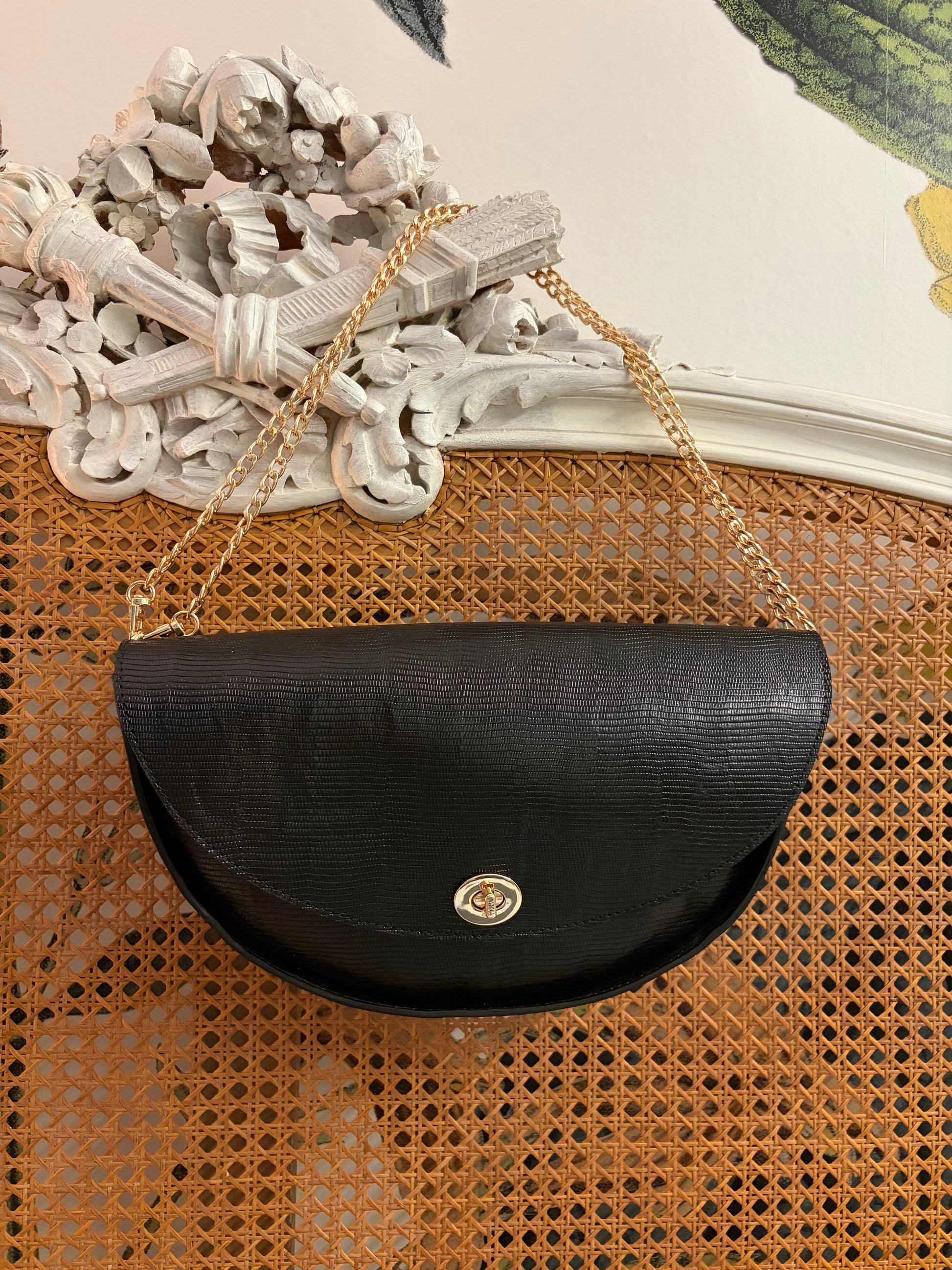 le sac est présenté de face accroché à un panneau en cannage. Il est en cuir noir texturé lézard et possède un fermoir et une anse en chainette dorés