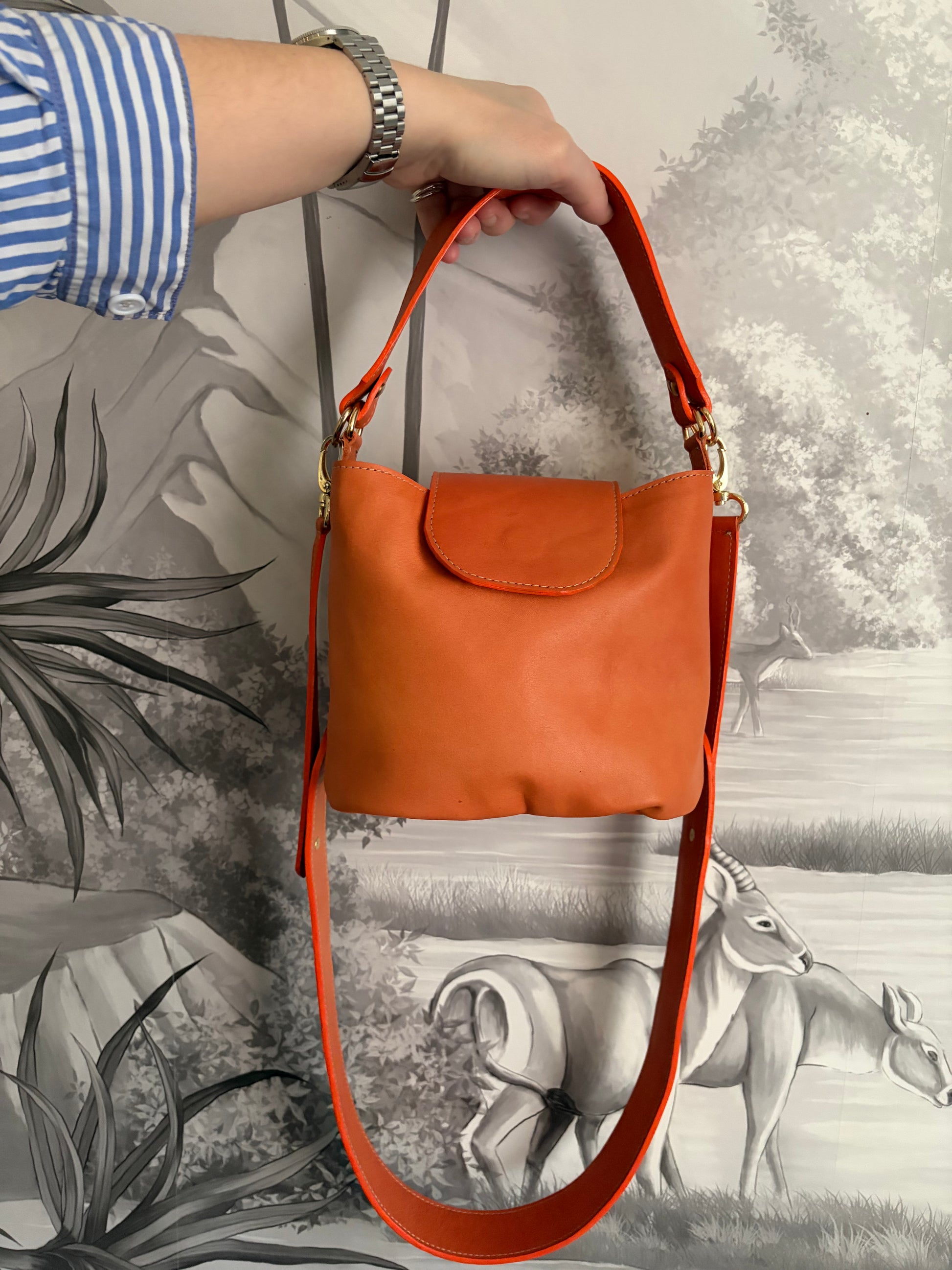 phot du sac seau Garance en cuir au tannage végétal orange porté à la main devant un mur avec un papier peint panoramique gris 