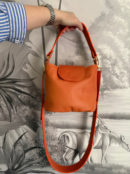 phot du sac seau Garance en cuir au tannage végétal orange porté à la main devant un mur avec un papier peint panoramique gris 