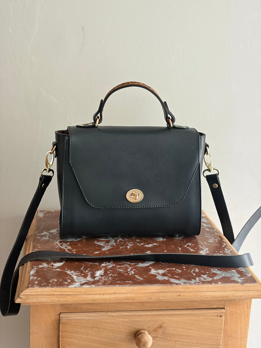 le sac simone en cuir noir est présenté posé sur une table en bois avec un plateau en marbre rouge. 
