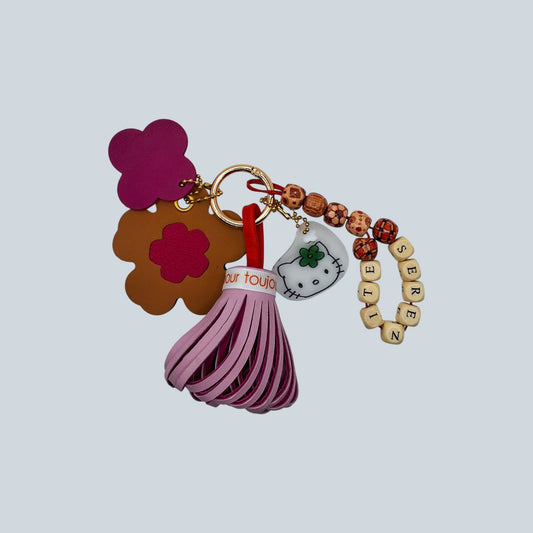 Bijou de sac avec cinq breloques en cuir et autres matériaux fixées à un mousqueton doré : un pompon bicolore rose clair et rose foncé ceinturé d’un ruban en gros grain marqué « amour toujours », une breloque en cuir marron et rose en forme de fleur, une breloque en forme de trèfle rose et rouge marquée M, une breloque en perles de bois avec motifs fantaisie formant le mot « SÉRÉNITÉ », et une breloque en polymère représentant une tête de chaton.