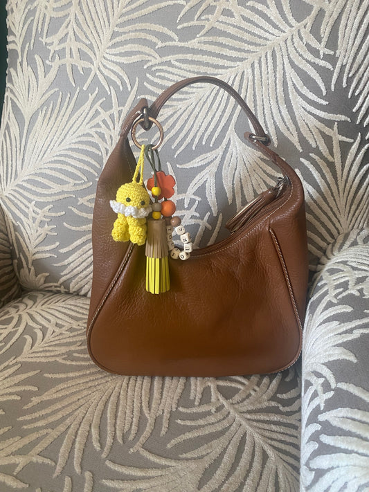 Bijou de sac en cuir avec trois breloques fixées à un mousqueton doré ovale : un pompon beige et jaune avec trois perles en bois, une breloque en perles de bois formant le mot « TOUJOURS », et une breloque en crochet faite à la main de couleur jaune.