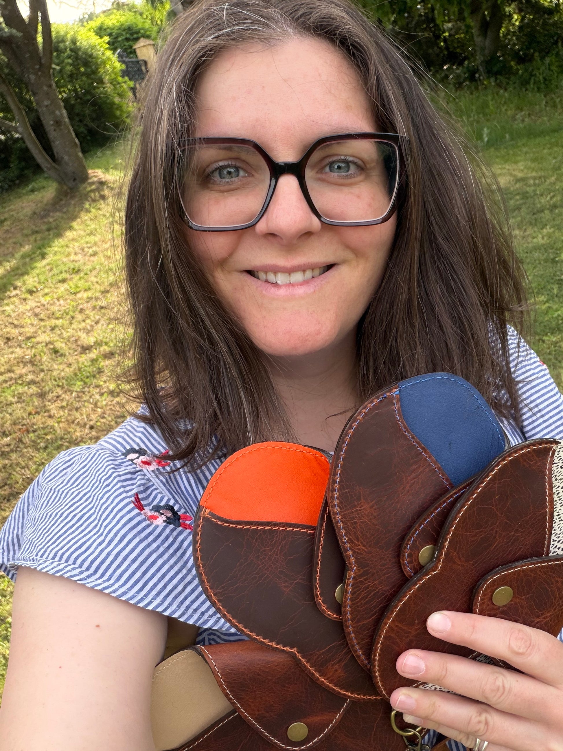 Une jeune femme souriante tient dans sa main 4 étuis à lunettes dans un jardin